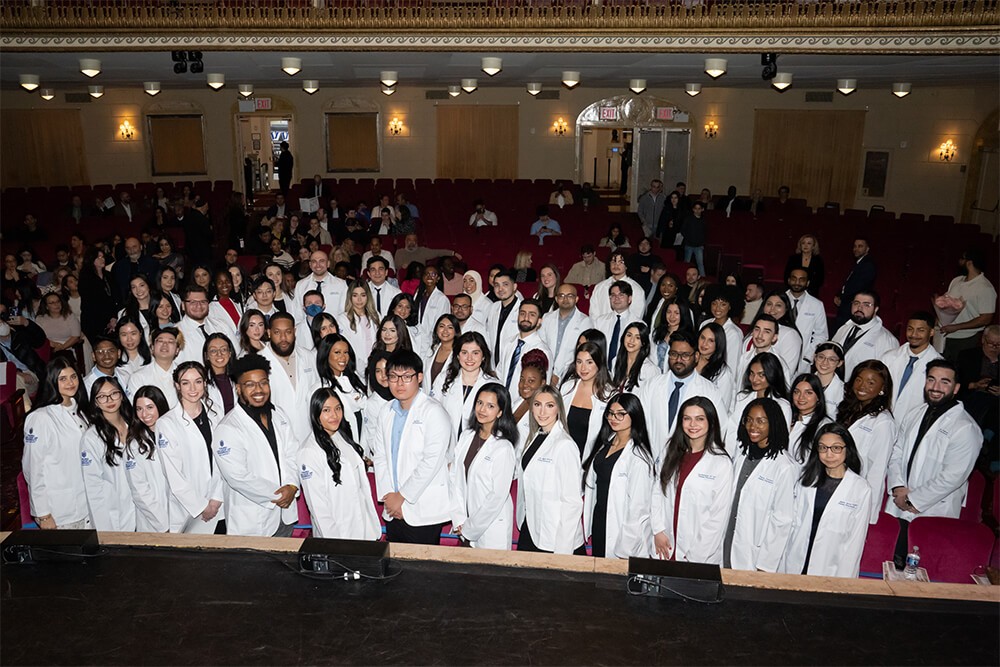 Seventy-two men and women lined up standing in several rows in auditorium behind one another smiling at camera in their new white coats.