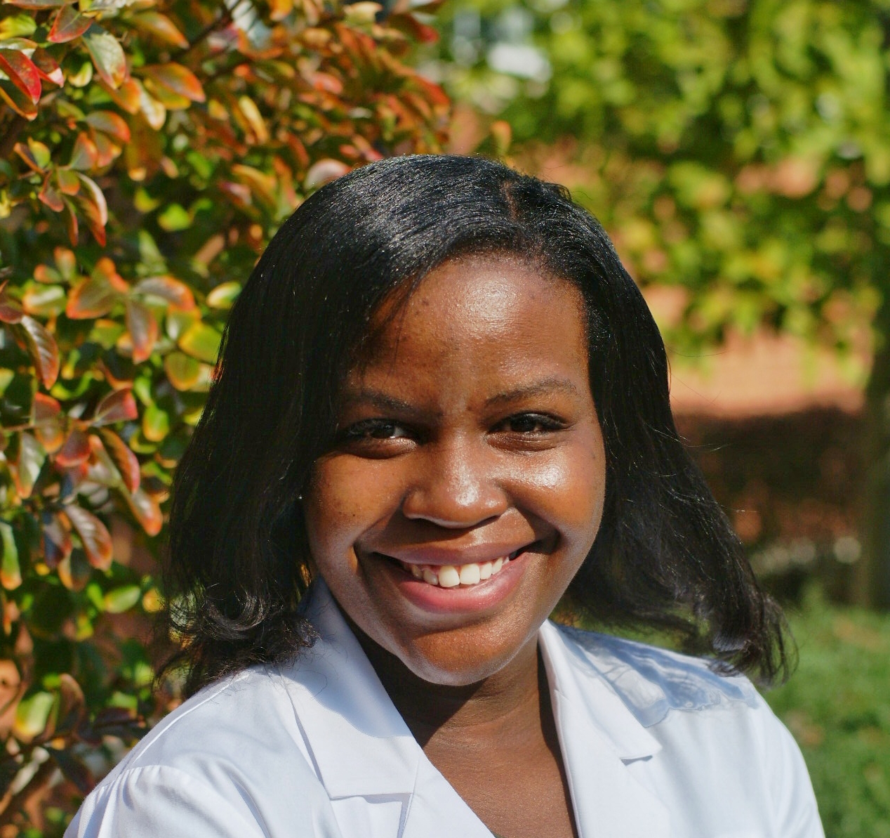 Lendelle Raymond, smiling outside wearing white coat, in garden surrounded by shrubbery 