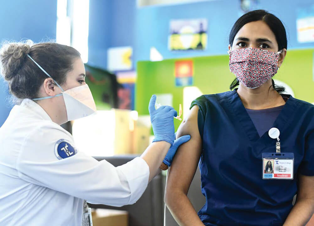Professor Rebecca Kavanagh, Pharm.D., wearing a mask, administers a vaccine to the right arm of a student who is sitting with her sleeve rolled up.
