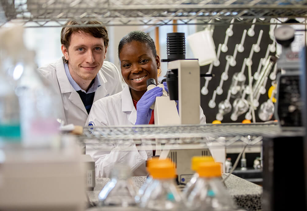 African American female pharmacy student in white coat and gloves holding microscope with white male pharmacy student in white coat next to her