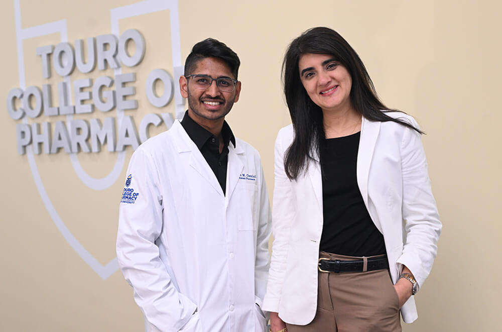 Associate dean of curriculum Batoul Senhaji-Tomza (right) with pharmacy student Alec Chunilall (left) in white coats standing to the right of a Touro College of Pharmacy sign on the wall